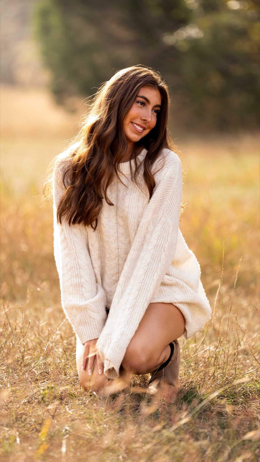 A senior portrait in a sweater kneeling in a golden hour field